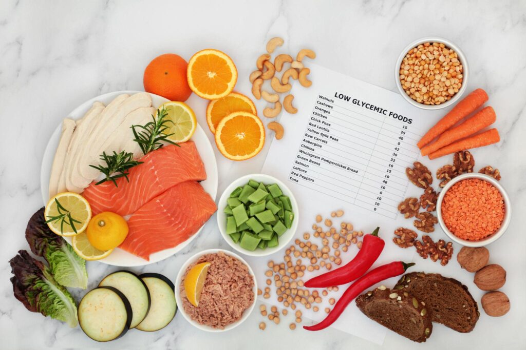 A collection of low-GI ingredients spread across a marble countertop, showing fresh fish, lean poultry, whole grain bread, nuts, seeds, and citrus fruit next to a glycemic index reference chart.